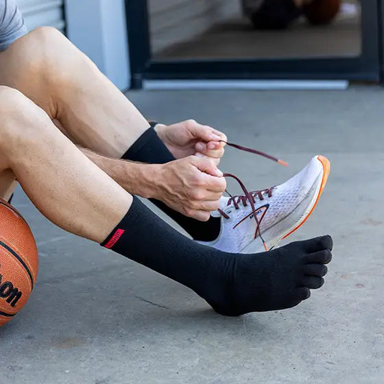 Person som sitter på marken med en basketboll, iklädd svarta tåstrumpor från Injinji och håller på att knyta sina löparskor med orange sula. Fokus på fotens naturliga rörelsefrihet med tåseparerande design