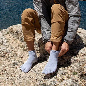 Person sitter på en klippa vid vattnet iklädd beige friluftsbyxor och grå tåstrumpor från Injinji