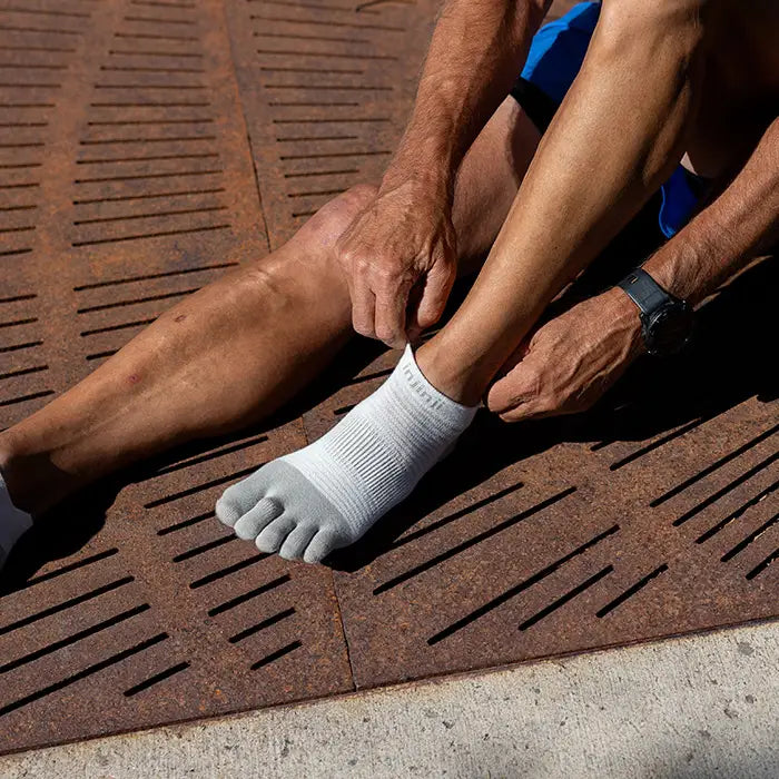 Person sitter på marken och justerar sina sandfärgade Injinji tåstrumpor i solljus, med ett ben utsträckt och det andra böjt.