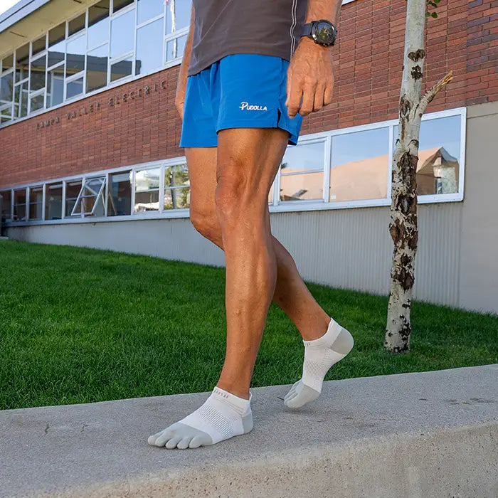 Man iförd grå tåstrumpor från Injinji står på en betongkant utomhus framför en tegelbyggnad, klädd i blå löparshorts.