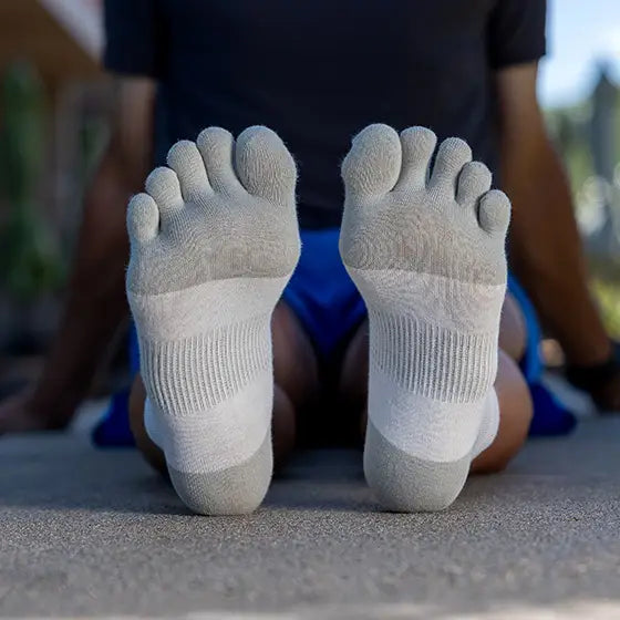 Person sitter på marken med benen utsträckta och visar undersidan av sina ljusgrå Injinji tåstrumpor i sandfärgad ton.