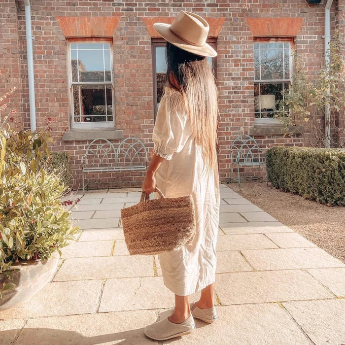 Kvinna i ljus sommarklänning, halmhatt och slip-on skor från VIBAe står utomhus med en flätad korgväska i solen framför ett tegelhus.