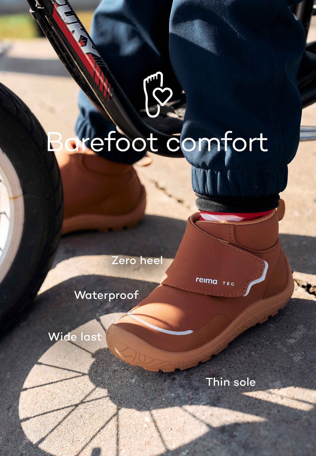 Närbild på ett barn ifört barfotainspirerade vattentäta skor från Reima TEC i brun färg, sittandes på en balanscykel. Text i bilden beskriver egenskaperna: zero heel, waterproof, wide last och thin sole – med budskapet "Barefoot comfort".