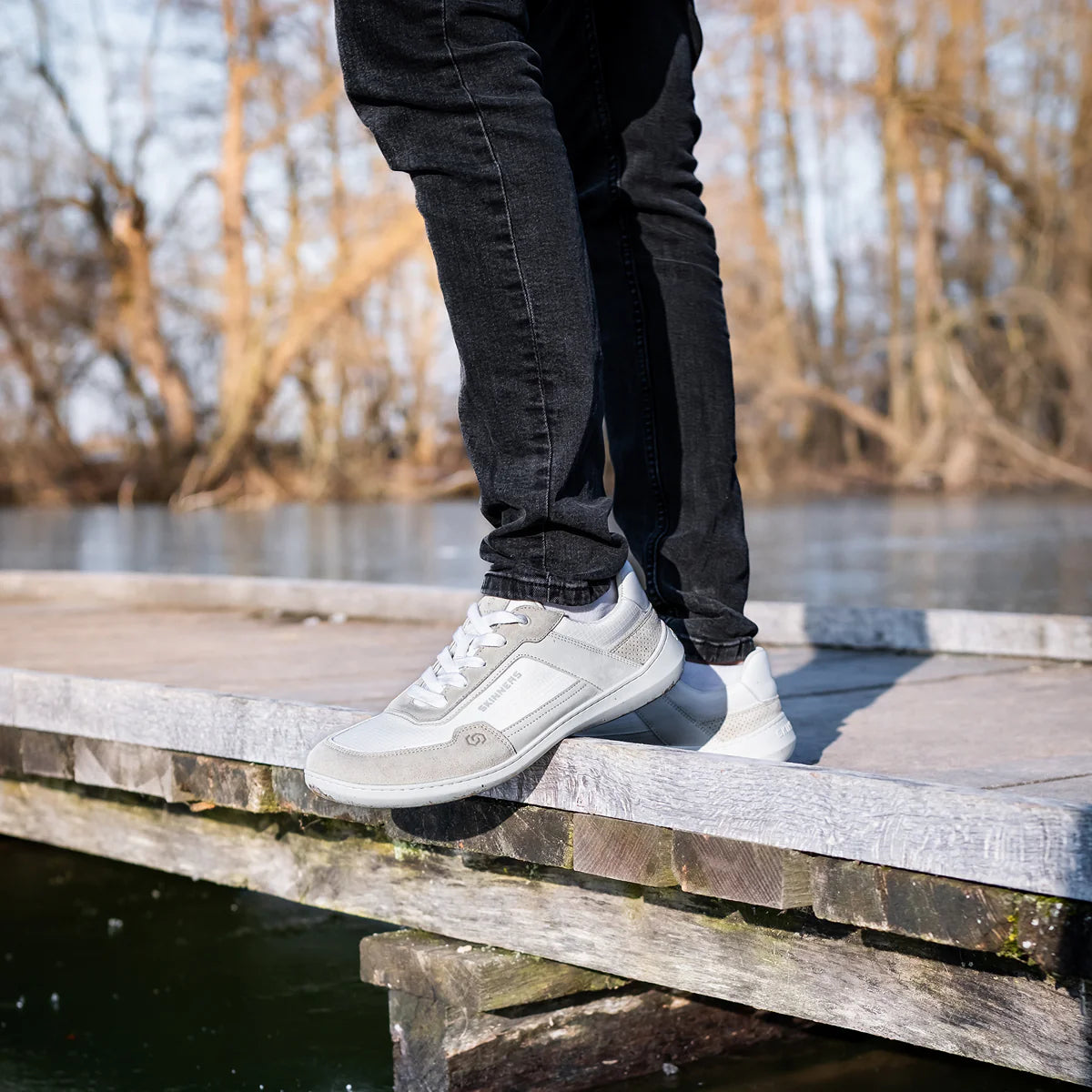 Person står på en brygga iförd Skinners Allrounder White barfotaskor – minimalistiska sneakers i vitt och ljusgrått med bred tåbox, perfekt för naturlig rörelse året runt.