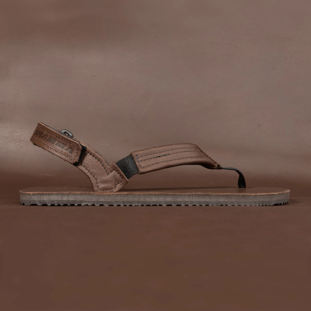 Shamma Sandals Pacific Avenue – minimalistisk barfotasko i brunt läder visad i profil mot brun bakgrund. Tydlig vy över fotremmar, vadderad vristrem och den tunna Vibram-sulan med låg profil för naturlig markkontakt