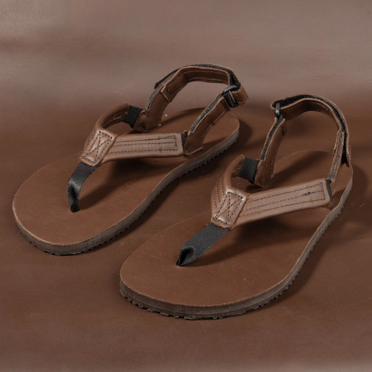 Shamma Sandals Pacific Avenue – par barfotaskor i brunt läder fotograferade snett ovanifrån mot brun bakgrund. Fokus på den breda tåremmen, justerbara hälremmar och den tunna, plana sulan med greppvänligt mönster.