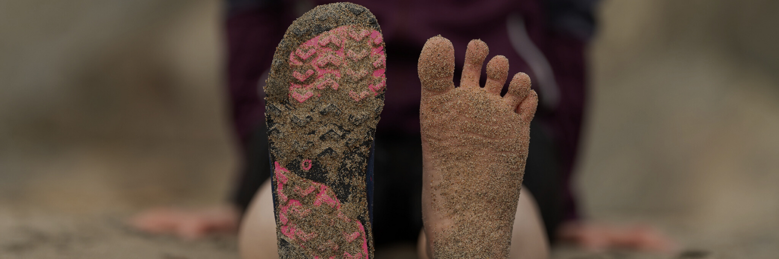 Närbild på en barfotasko och en bar fot täckt av sand på stranden. Skons sula har rosa mönster och visar kontrasten mellan barfotakänsla och skyddad fot.