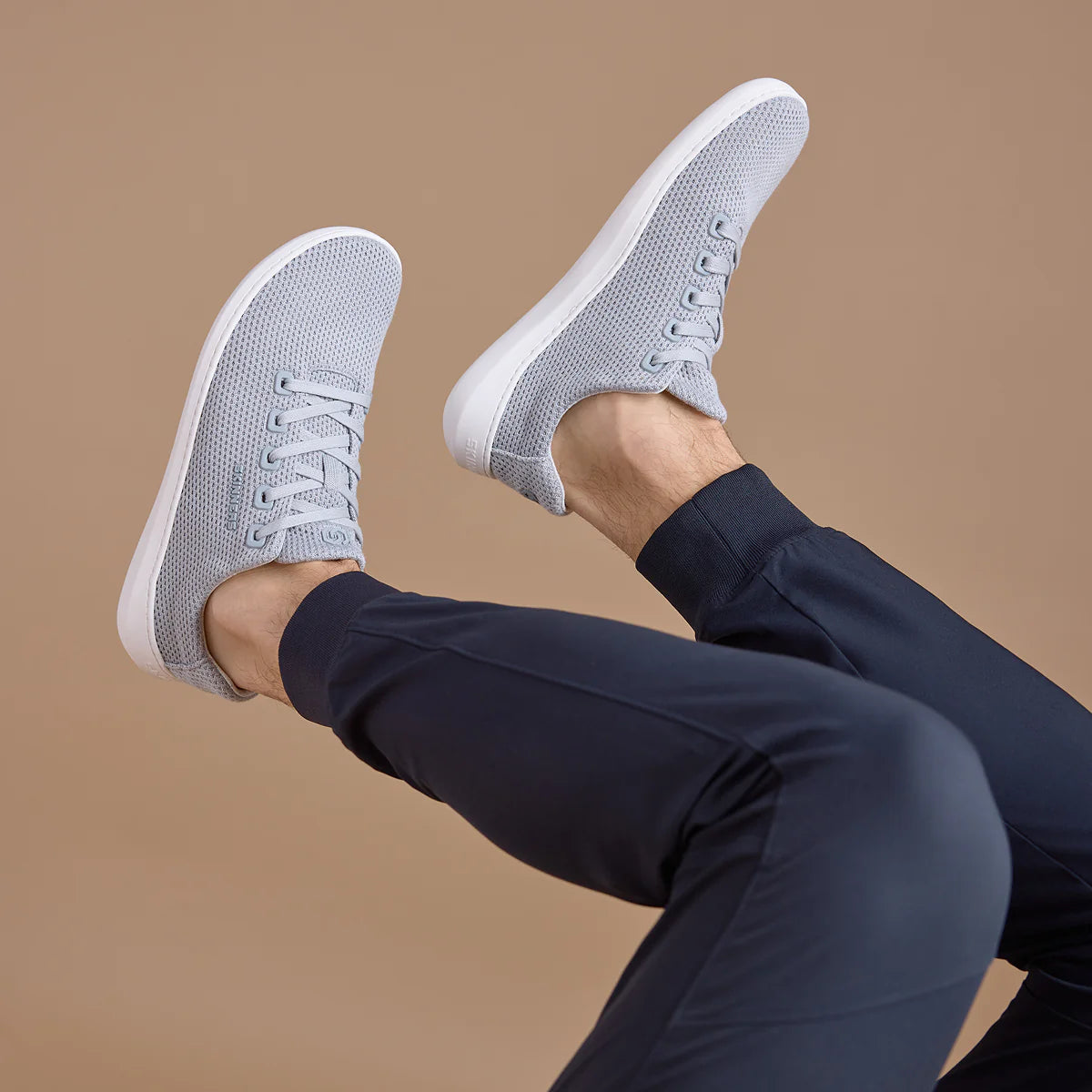 Person iförd Skinners Globetrotter barfotaskor i färgen Cloudy Gray med vit sula. Skorna bärs till mörkblå byxor och benen är upplyfta mot en beige bakgrund. Skons lätta meshmaterial och minimalistiska design framhävs