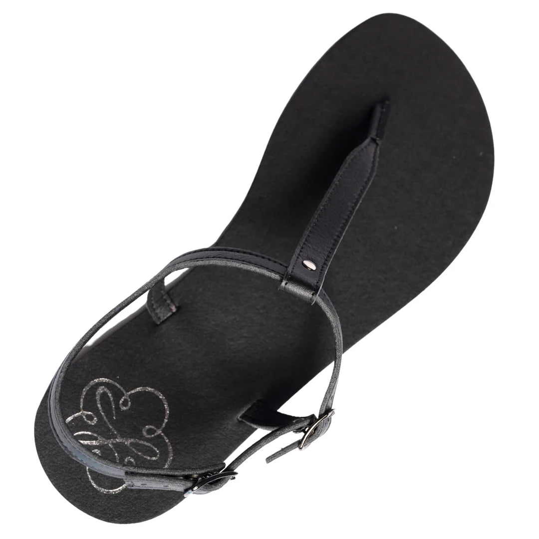 Produktbild på Lonnies sandaler Lux Little Black sedd uppifrån. Den minimalistiska barfotaskon har svarta remmar i T-design, ett justerbart spänne och en slät svart sula med Lonnies logotyp tryckt i hälen. Designad för barfotakänsla med stilren komfort.