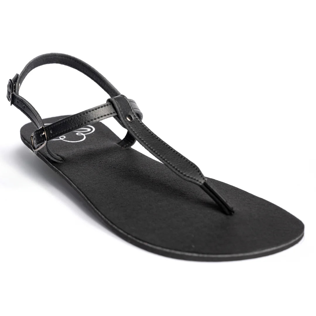 Produktbild på Lonnies barfotasko i modellen Lux Little Black, fotograferad i sned vinkel framifrån. Sandalen har tunna svarta remmar med spänne, T-remdesign och en minimalistisk svart sula. Perfekt för barfotakänsla med stilren look