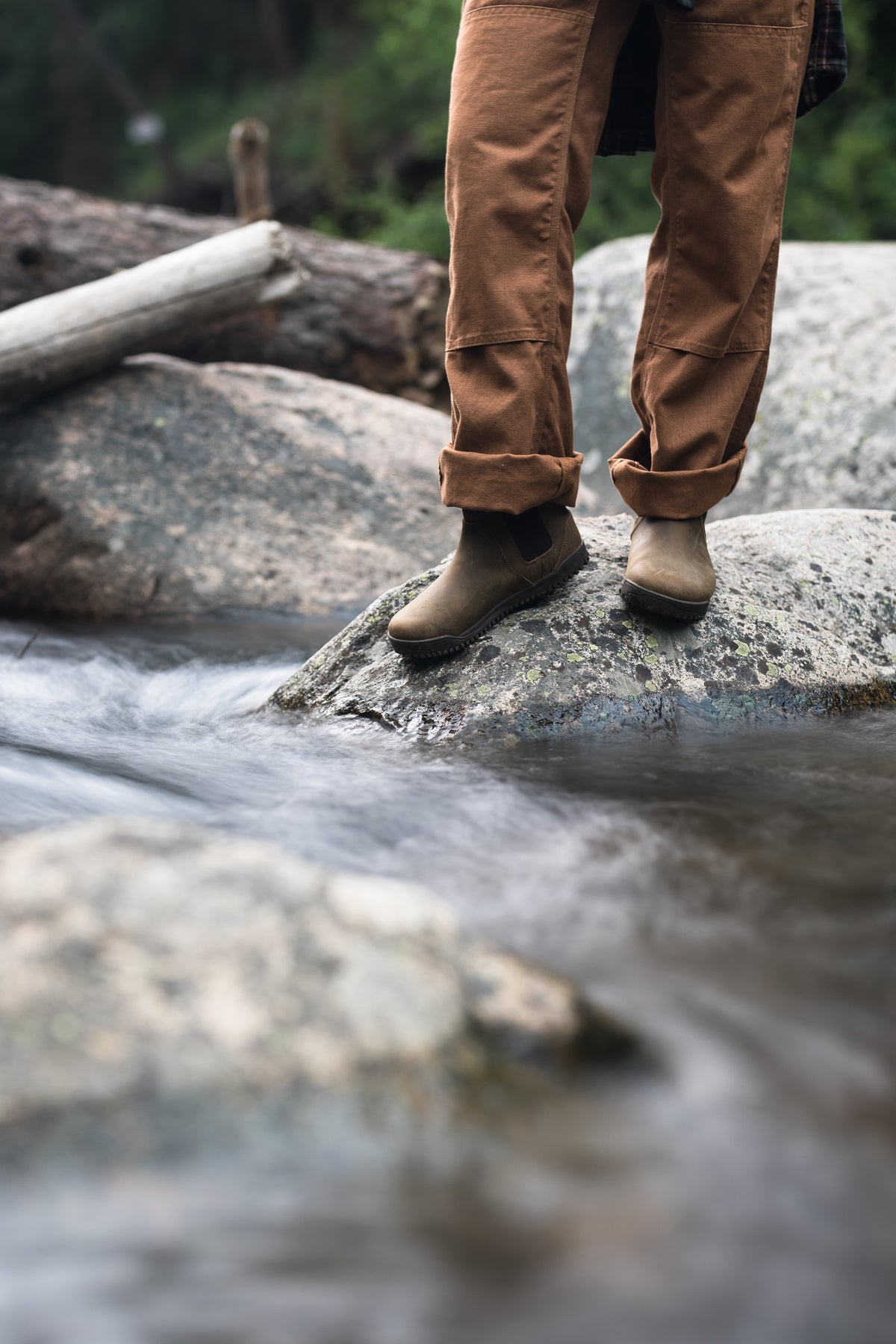 Person står på en sten i ett strömmande vattendrag, iförd bruna Ridgeway Chelsea-kängor från Xero Shoes och uppvikta arbetsbyxor i samma ton. Bilden är tagen i en naturlig miljö med rinnande vatten, stenblock och skog – och signalerar robust barfotakomfort i terräng.