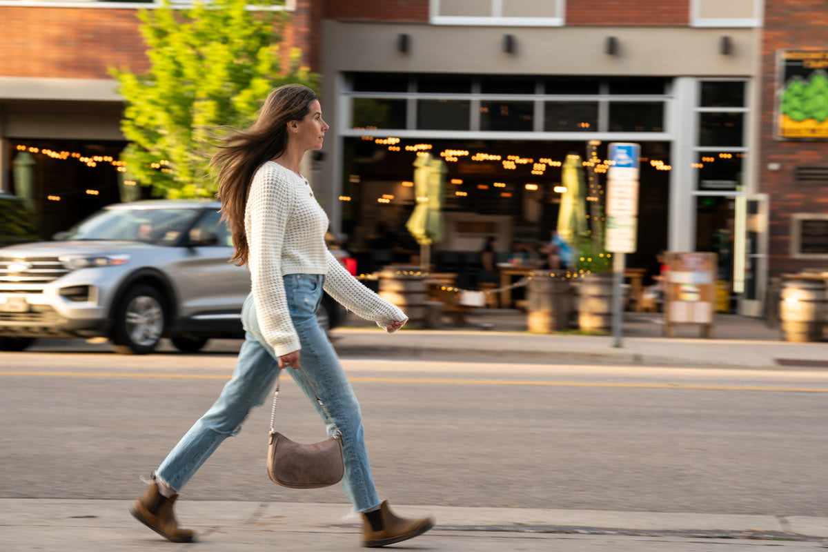 Kvinna promenerar tvärs över en stadsgata i kvällssol, iförd blå jeans och bruna Ridgeway Chelsea-kängor från Xero Shoes. I bakgrunden syns en uteservering med ljusslingor och ett stadsliv i rörelse.