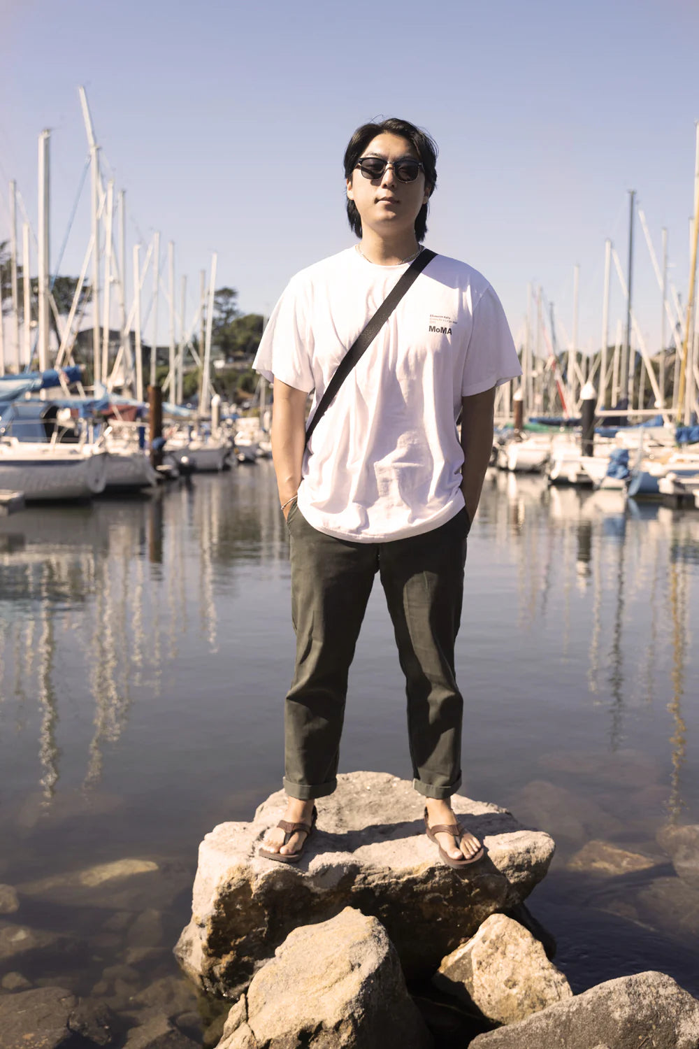 Person står barfota i ett par Shamma Sandals Pacific Avenue på en klippa vid en småbåtshamn. Sandalerna i brunt läder syns tydligt mot det marina bakgrunden med segelbåtar, vilket lyfter fram skons minimalistiska och somriga design