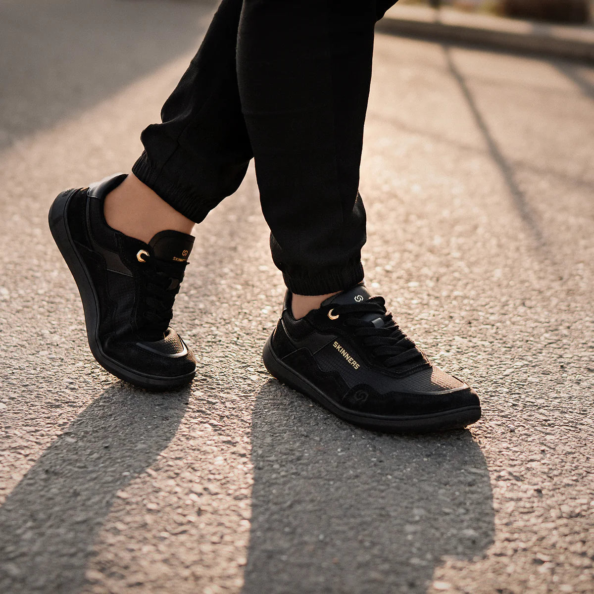 Person går på asfalt i minimalistiska barfotaskor Skinners Camina Black – svart design med gulddetaljer, låg sula och bred tåbox för naturlig rörelse och komfort.