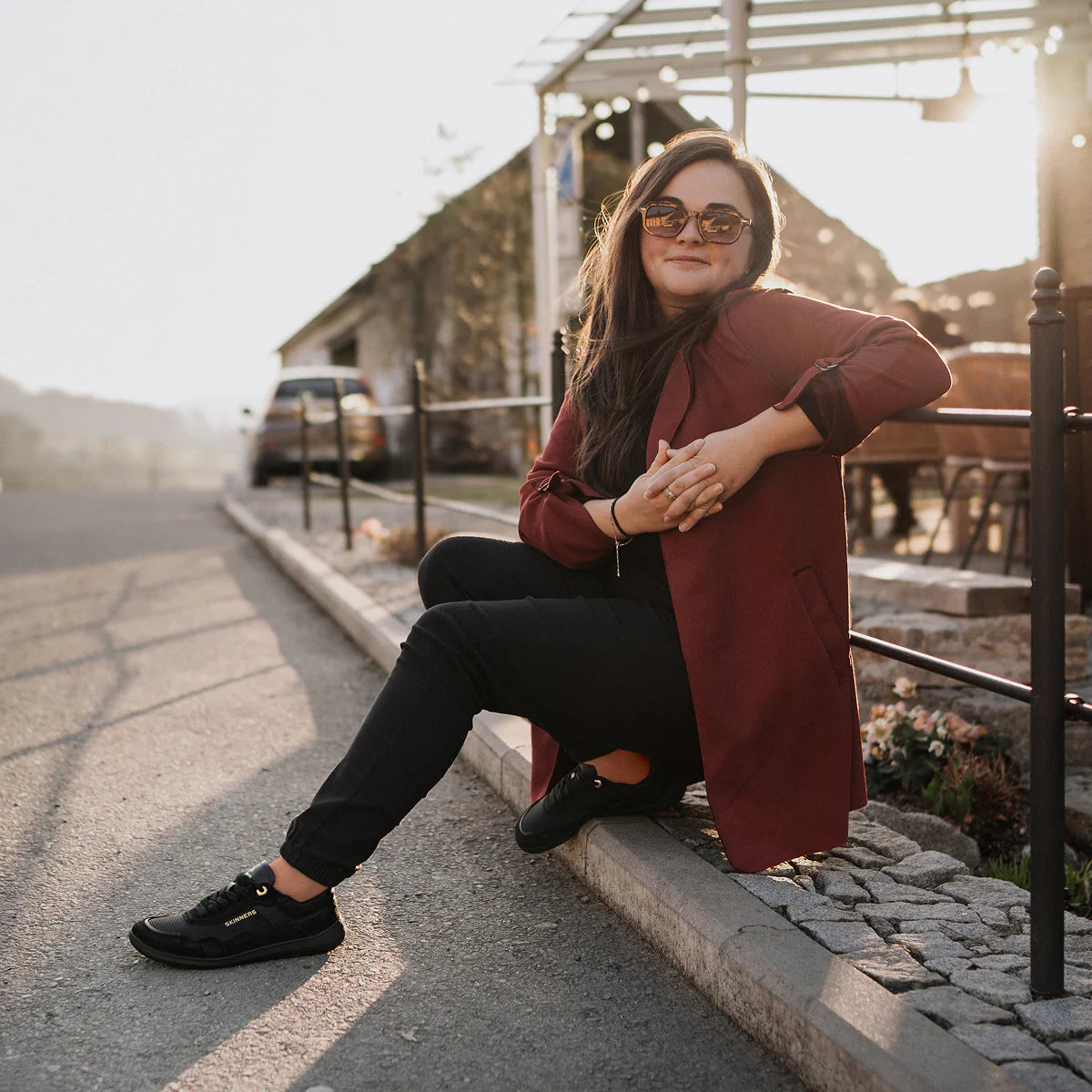 Kvinna i solglasögon och vinröd kappa sitter avslappnat på en trottoarkant i kvällssol, iförd minimalistiska barfotaskor Skinners Camina Black – svarta sneakers med bred tåbox och barfotakänsla.
