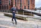 Man promenerar längs en kaj i stadsmiljö, iförd svarta Addict Road-barfotaskor från Joe Nimble, jeans och en grå långärmad tröja. I bakgrunden syns röda tegelbyggnader och vatten.