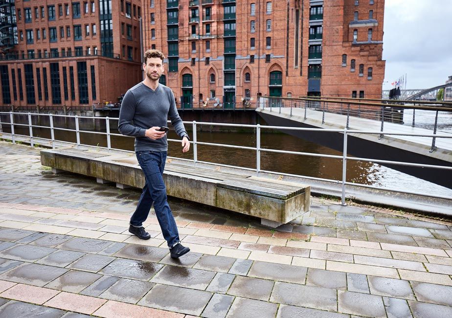 Man promenerar längs en kaj i stadsmiljö, iförd svarta Addict Road-barfotaskor från Joe Nimble, jeans och en grå långärmad tröja. I bakgrunden syns röda tegelbyggnader och vatten.
