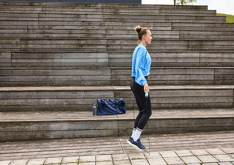 Kvinna tränar utomhus genom att hoppa hopprep på trätrappor, iförd blå Addict Road-barfotaskor från Joe Nimble, svarta tights och en blå tröja. En blå sportväska och vattenflaska står bakom henne – bilden förmedlar rörelsefrihet och naturlig dämpning i stadsmiljö.
