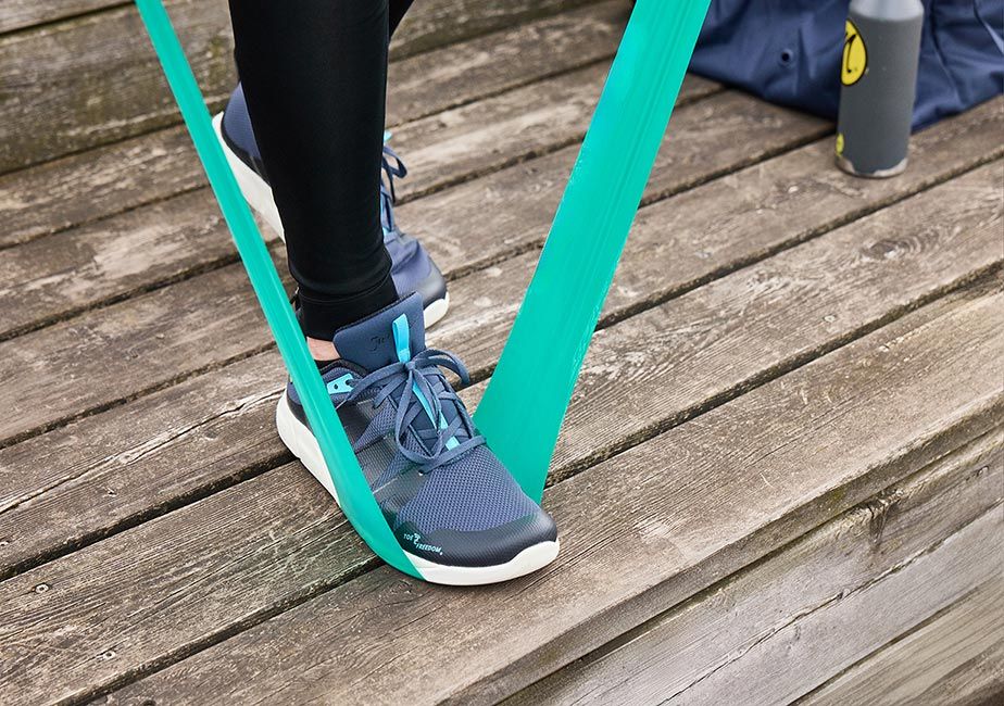 Närbild på blå Addict Road-skor från Joe Nimble under motståndsträning med ett turkost träningsband. Personen står på ett trädäck iklädd svarta tights; i bakgrunden syns en vattenflaska och träningsväska.