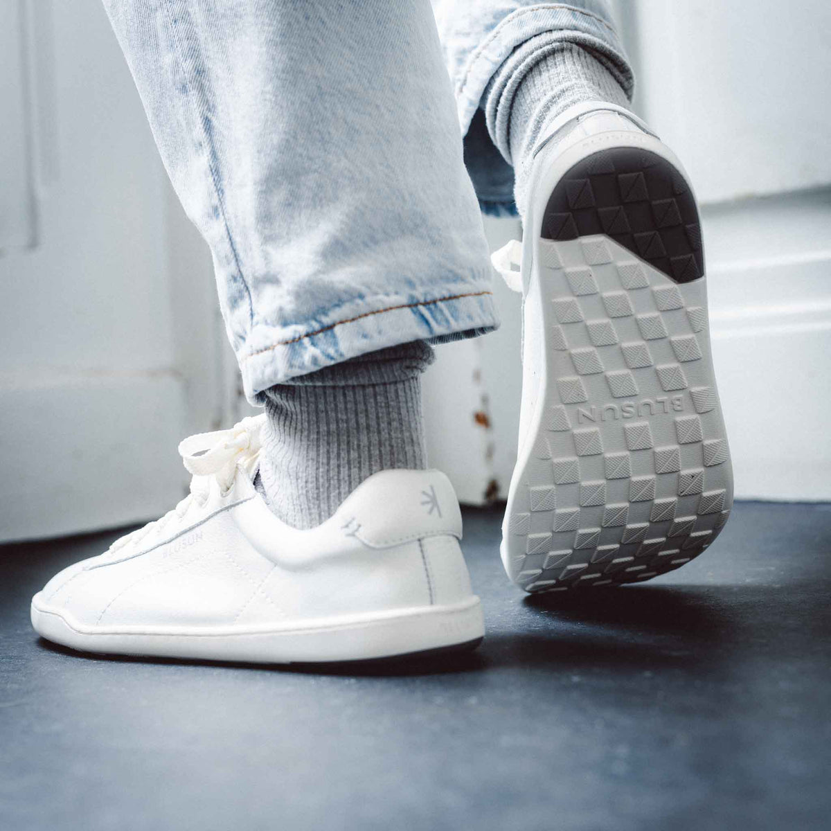 Person med ljusa jeans och gråa strumpor bär vita barfotaskor från Blusun. Fokus på skornas flexibla yttersula med greppmönster och märkets logotyp synlig i hälen.