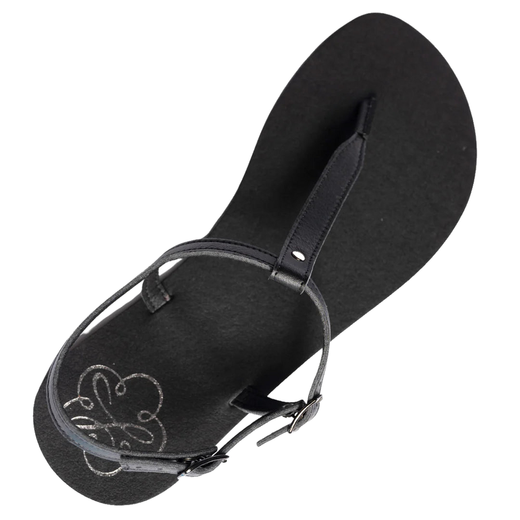 Produktbild på Lonnies sandaler Lux Little Black sedd uppifrån. Den minimalistiska barfotaskon har svarta remmar i T-design, ett justerbart spänne och en slät svart sula med Lonnies logotyp tryckt i hälen. Designad för barfotakänsla med stilren komfort.