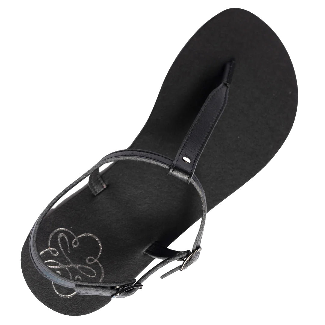 Produktbild på Lonnies sandaler Lux Little Black sedd uppifrån. Den minimalistiska barfotaskon har svarta remmar i T-design, ett justerbart spänne och en slät svart sula med Lonnies logotyp tryckt i hälen. Designad för barfotakänsla med stilren komfort.
