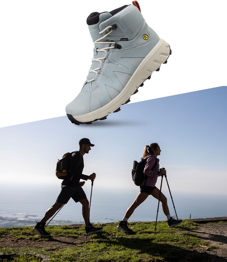 Produktbild av den ljusblå vandringskängan Mountain Addict från Joe Nimble med Vibram-sula, kombinerad med en siluettbild av två vandrare med stavar som går längs en havskant i morgonljus. 
