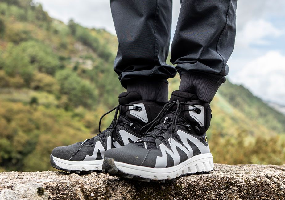 Närbild på ett par svarta Mountain Addict barfotaskor från Joe Nimble, burna av en person i svarta friluftsbyxor som står på en sten i grön bergsmiljö. Skorna har vita sidor och rejäl sula.