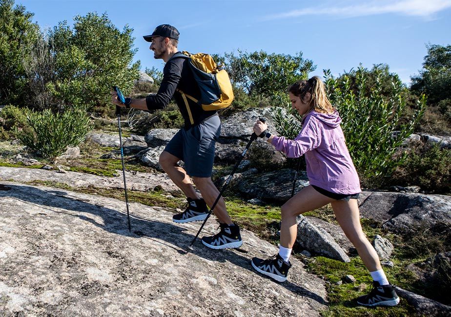 Två personer vandrar uppför en stenig sluttning i dagsljus, iförda svarta Joe Nimble Mountain Addict-barfotaskor med vita detaljer. Den ena bär shorts och ryggsäck, den andra en lila vindjacka – båda använder vandringsstavar. 
