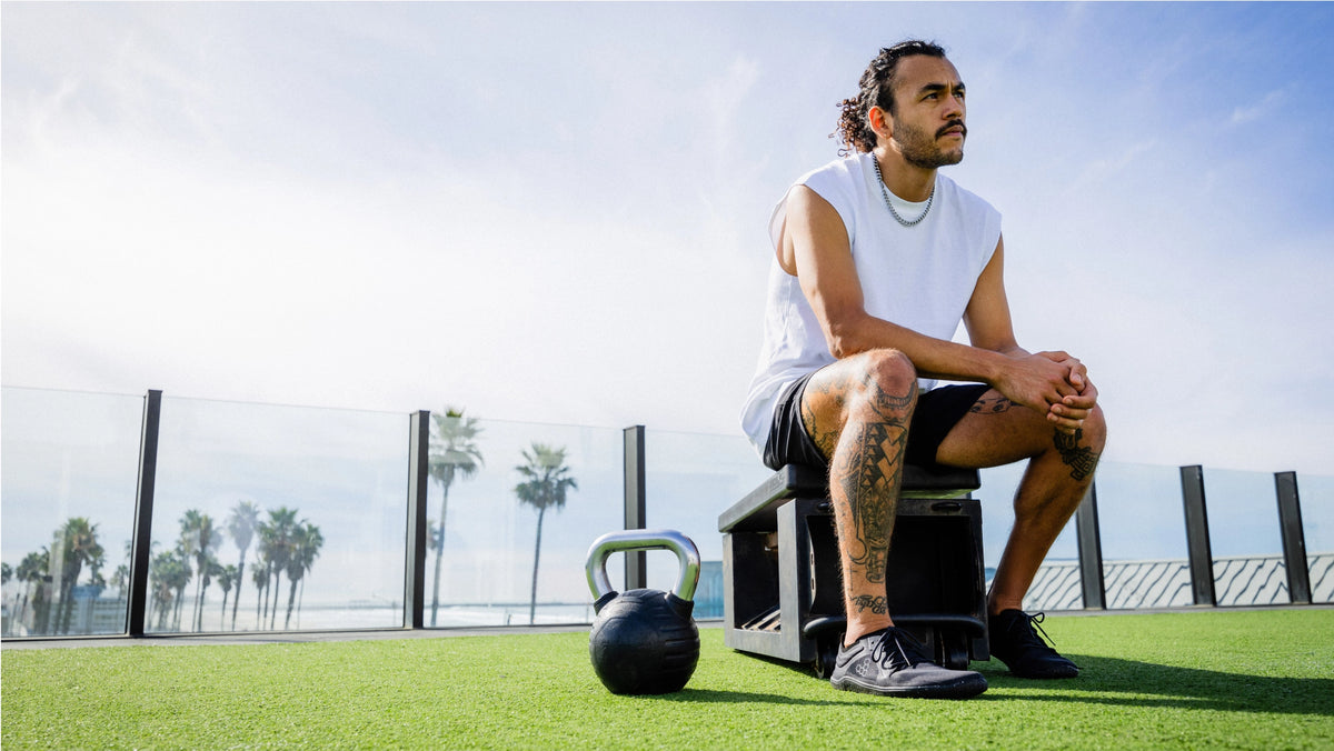 Man sitter på en träningsbänk på ett takgym med utsikt över palmer, iförd träningskläder och barfotaskon Primus Lite III i färgen Obsidian från Vivo Barefoot. En kettlebell står bredvid.